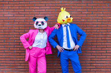 Man And Woman Wearing Vibrant Suits And Animal Masks Posing Together In Front Of Brick Wall