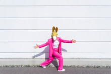 Woman Wearing Vibrant Pink Suit And Donkey Mask Posing In Front Of White Wall With Open Arms