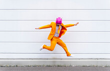 Man Wearing Vibrant Orange Suit And Hippo Mask Jumping In Front Of White Wall