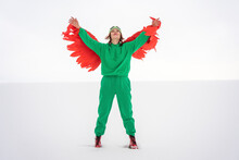 Carefree Woman In Green Bird Costume Raising Arms While Standing On Snow Against Sky