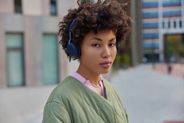 Wall Mural - Photo of serious good looking woman with Afro hair listens music in headphones enjoys audio player dressed in casual jacket uses wireless headphones poses against blurred background outside.