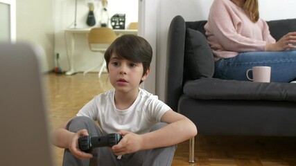 Wall Mural - boy playing video games at home. mother sitting on a sofa in background