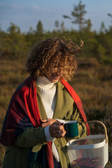 Wall Mural - Young curly woman warming up after collecting cranberries on swamp holding mug of hot tea or coffee. Happy smiling female in trendy trench enjoy spending weekend alone in countryside. Autumn leisure