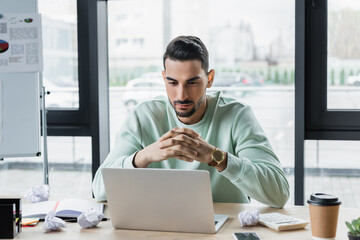 Wall Mural - Arabian businessman looking at laptop near coffee to go and crumpled paper on table