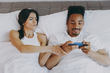 Wall Mural - Top view sad young couple two friends family in casual white clothes lying in bed woman look angry at man playing game at mobile cell phone spend time together in bedroom lounge home own room house