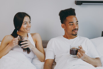 Wall Mural - Happy young couple two man woman family in casual white clothes sit in bed hold transparent cup drink coffee tea look aside relax spend time together in bedroom lounge home house wake up good day