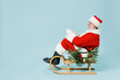 © ViDi Studio - Full size side view old Santa Claus man 50s in Christmas hat red suit clothes sitting in sled isolated on plain blue background studio. Happy New Year 2022 celebration merry ho x-mas holiday concept.