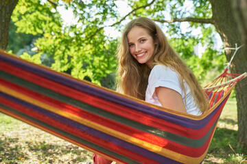 Horizontal image of a young woman laying in hammock relaxing in the park. Pretty female resting on hammock hanging between trees smiling and looking at camera in a forest.