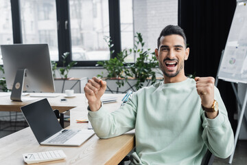 Wall Mural - Excited muslim businessman showing yes gesture near calculator and laptop