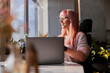Pink haired woman with elegant glasses works on laptop near window at home