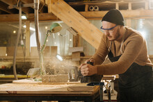 Keen Carpenter In Watch Cap Shaving Wood With Electric Plane In A Big Workshop.