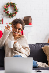 Wall Mural - Smiling african american woman with cup having video call on laptop during christmas