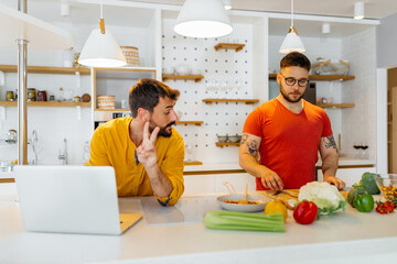 Wall Mural - A gay couple cooking vegan dinner at home. A man in the yellow shirt looking at a laptop and trying to follow a recipe while his husband chopping fresh vegetables.