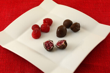 Wall Mural - Frozen raspberries coated in white chocolate and dark chocolate with fresh raspberries on white snack plate on red tablecloth