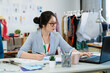 © PR Image Factory - charming female entrepreneur sitting at work desk with garment equipments and analyzing information from laptop computer. concentrated asian woman fashion designer using notebook pc creative idea.
