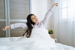 © PR Image Factory - Woman stretching in bedroom after wake up. young asian korean beautiful lady sitting in comfort white bed smiling enjoy morning with sunlight. happy charming female raised arms and hands joyful face.