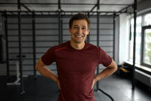 Indoor Portrait Of Cheerful Young Handsome Caucasian Man In Red Sport T-shirt Posing Against Gym, Smiling Holding Hands On Waist, Resting During Fitness Workout. Active Lifestyle. Fitness
