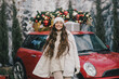 © polinaloves - Happy beautiful woman wearing knitted sweater and woolen hat standing near red car with Christmas tree on the top under snowfall.