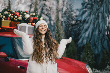 Happy Beautiful Woman Wearing Knitted Sweater And Woolen Hat Standing Near Red Car With Christmas Tree On The Top Under Snowfall.