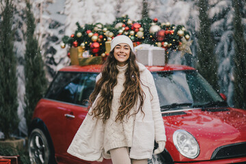 Happy beautiful woman wearing knitted sweater and woolen hat standing near red car with Christmas tree on the top under snowfall.