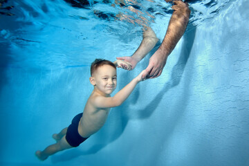 Wall Mural - A little boy swims under the water in the pool. Dad puts his hands under the water and catches the baby. Portrait. Close-up. Concept. Horizontal orientation.