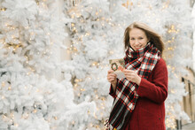 Portrait Of Smiling Young Woman Standing On The Street With Christmas Card In Front Of White Christmas Trees