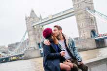 Two Young Tourists Sitting On Wall, Using Smartphone, With  London Bridge In Background