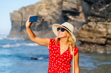 Blond Woman Wearing Red Dress And Hat And Using Smartphone And Taking A Selfie At The Beach