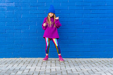 Girl Wearing Blue Cap And Oversized Pink Pullover Standing In Front Of Blue Wall Dancing