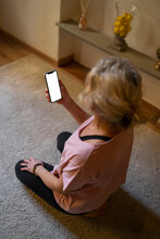 Mature Woman Sitting On Carpet In The Living Room Looking At Display Of Cell Phone