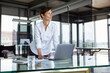 © Rainer Berg/Westend61 - Businesswoman standing at glass table in office with laptop looking sideways
