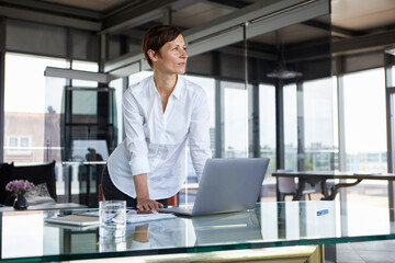 Businesswoman standing at glass table in office with laptop looking sideways