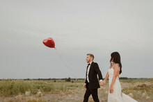 Bride And Groom With Heart Shape Balloon Holding Hands While Walking Against Clear Sky