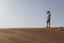 Male Tourist Shielding Eyes While Standing On Sand Dune Against Clear Sky At Dubai, United Arab Emirates
