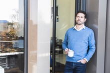 Businessman Holding Coffee Cup While Leaning On Wall With Hand In Pocket At Office Entrance