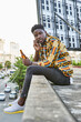 © victor espadas/Westend61 - Young man in patterned shirt sitting on roof terrace in the city talking on the phone, Maputo, Mozambique