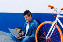 Smiling Mature Man Using Laptop By Orange Bicycle While Sitting Against Wall