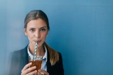 Portrait Of A Young Businesswoman Against Blue Background, Drinking Iced Tea With A Straw