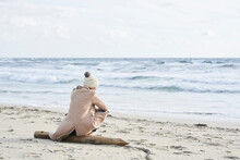 Spain, Menorca, back view of senior woman wearing bobble hat and coat sitting on the beach in winter