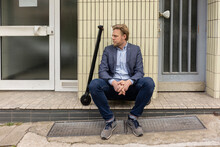 Businessman With E-Scooter Sitting On Step In Front Of A House