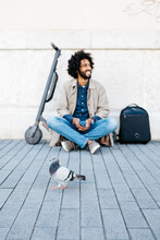 Smiling Man Sitting On His E-Scooter On Pavement While A Pigeon Passing By In The Foreground