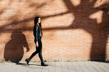 Young Woman With A Red Bag Walking In Front Of A Brick Wall And A Shadow Of Tree