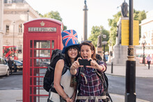 Cheerful Woman Taking Selfie With Friend Wearing British Flag Hat Against Red Telephone Box In City
