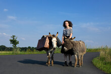 Young Woman Standing On Country Road With Donkeys Carrying Baggage