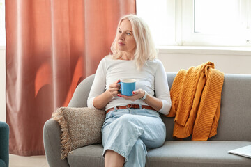 Sticker - Beautiful mature woman with cup of coffee sitting on sofa at home