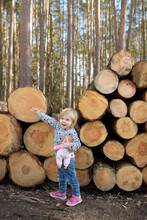 Portrait Of Happy Little Girl With Doll Standing In Front Of Stack Of Wood In The Forest