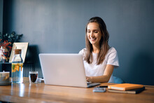 Smiling Young Woman Using Laptop On Desk Against Wall In Home Office