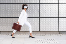 Young Businesswoman With Briefcase Using Virtual Reality Glasses While Walking Against Silver Wall