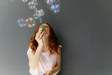 Redheaded Woman Leaning Against Grey Wall Blowing Soap Bubbles