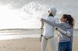 © MAGIC UNICORN/Westend61 - Smiling woman standing arm in arm with robot on the beach